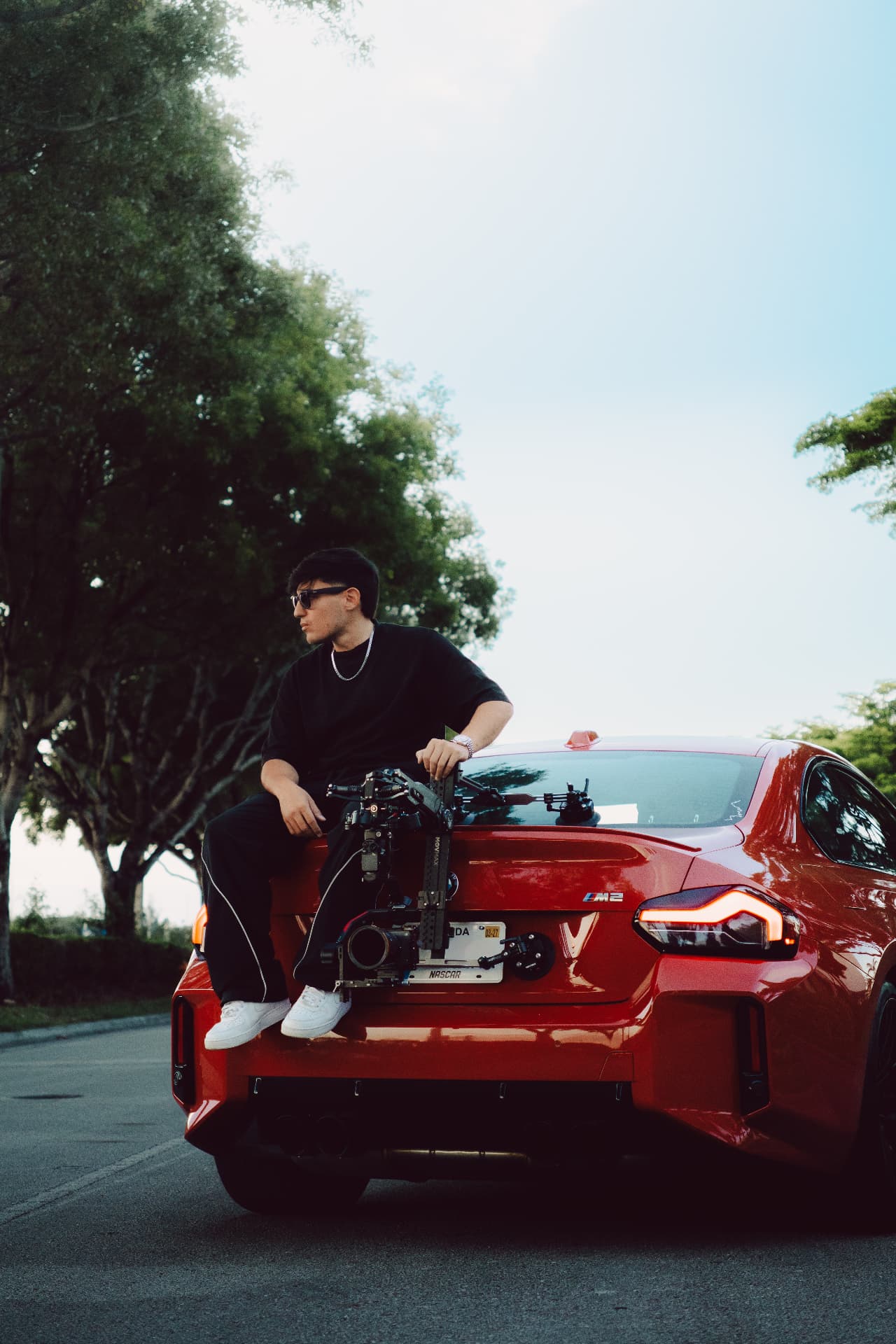 Andres sitting on the trunk of his red BMW M2 holding a camera rig
